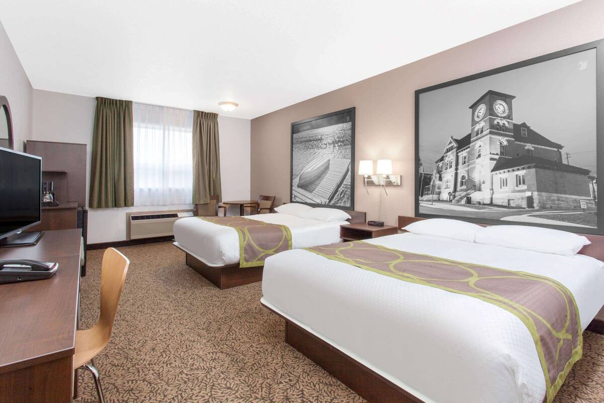 Habitación de hotel con dos camas, un escritorio con computadora, una silla, un televisor y un cuadro grande en la pared.