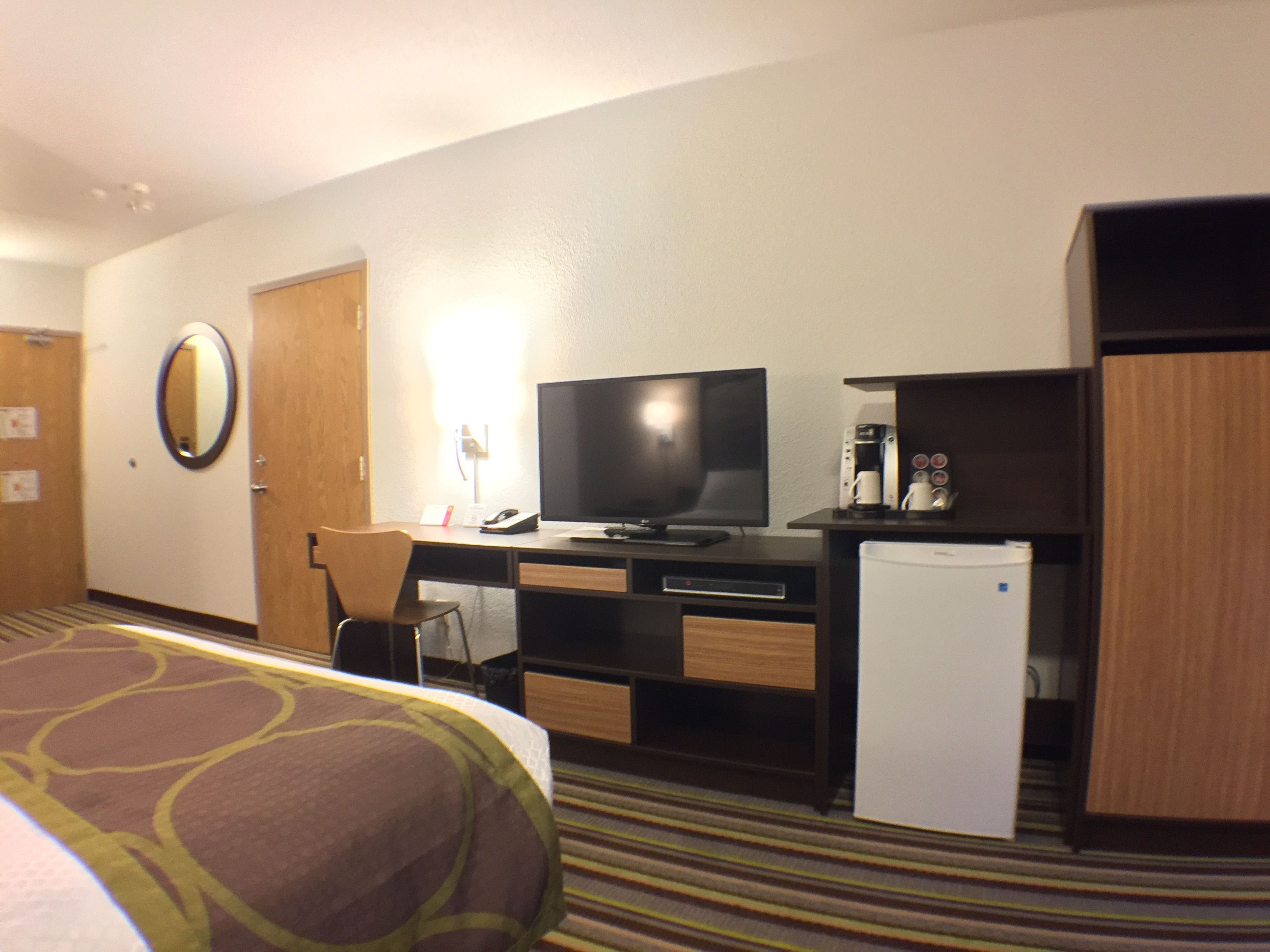 A hotel room with a bed, a desk with a television, a coffee machine, and a refrigerator.
