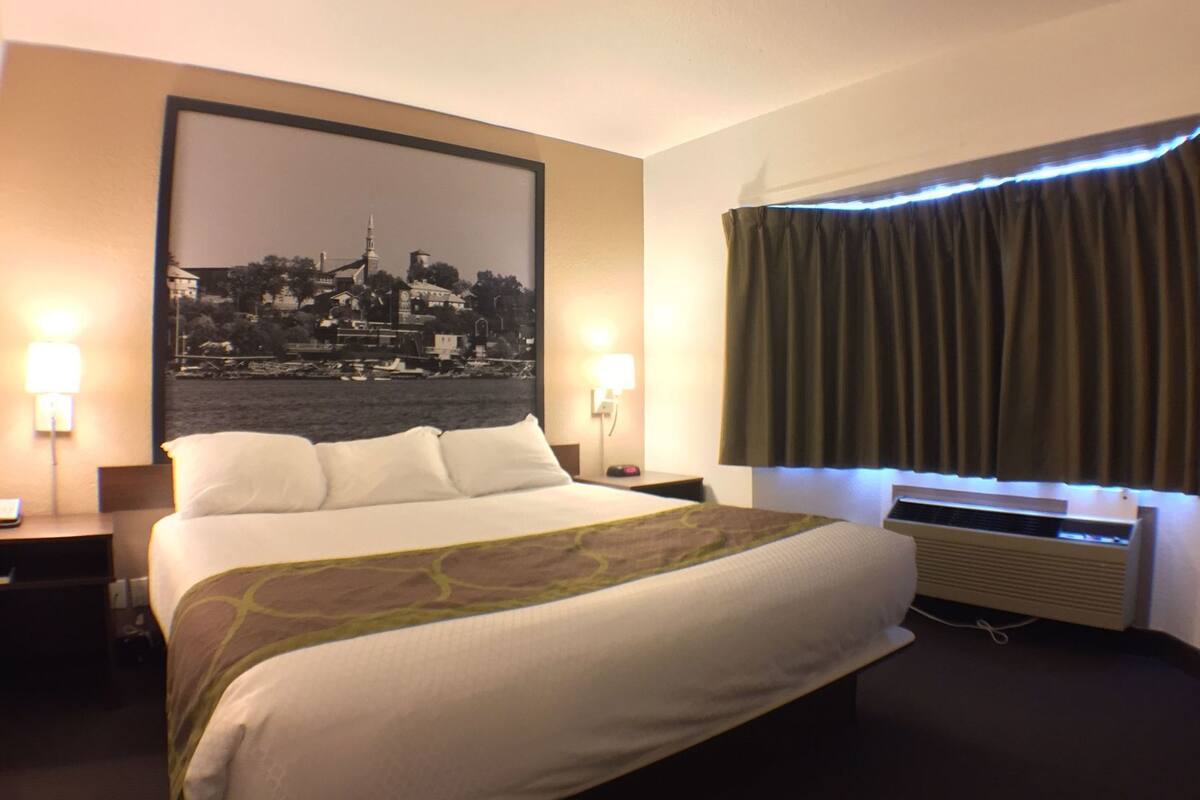 Habitación de hotel con cama, un cartel con un paisaje urbano, una ventana con cortinas y un aire acondicionado instalado en la pared.