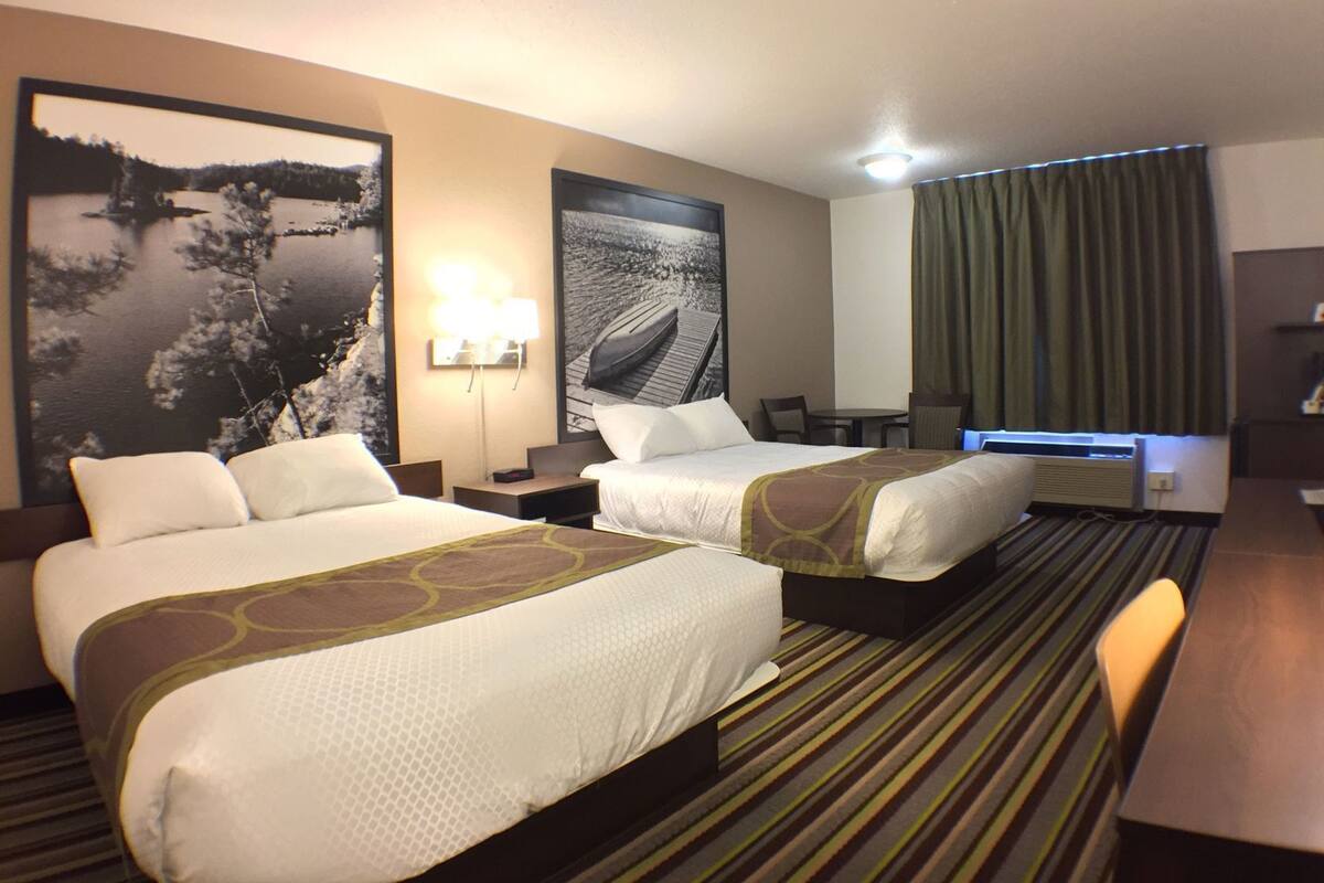Habitación de hotel con dos camas, un escritorio, una silla, una lámpara y un ventanal grande con cortinas.