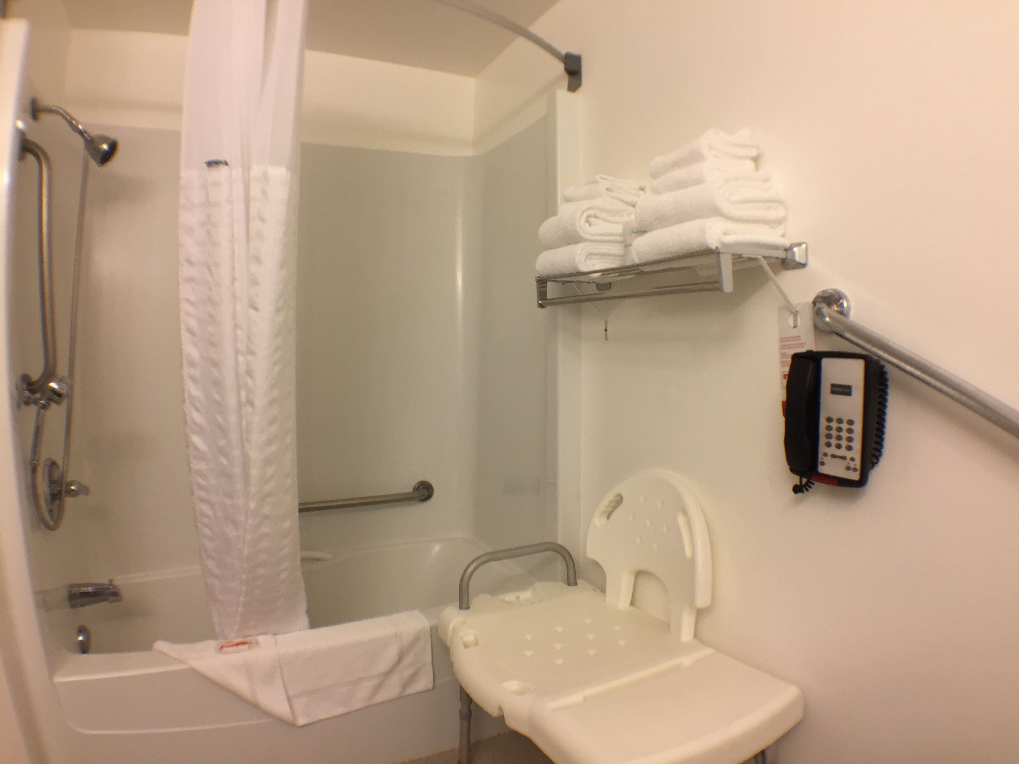 Un baño con ducha, inodoro, barra de apoyo, botón de asistencia y estante con toallas dobladas.