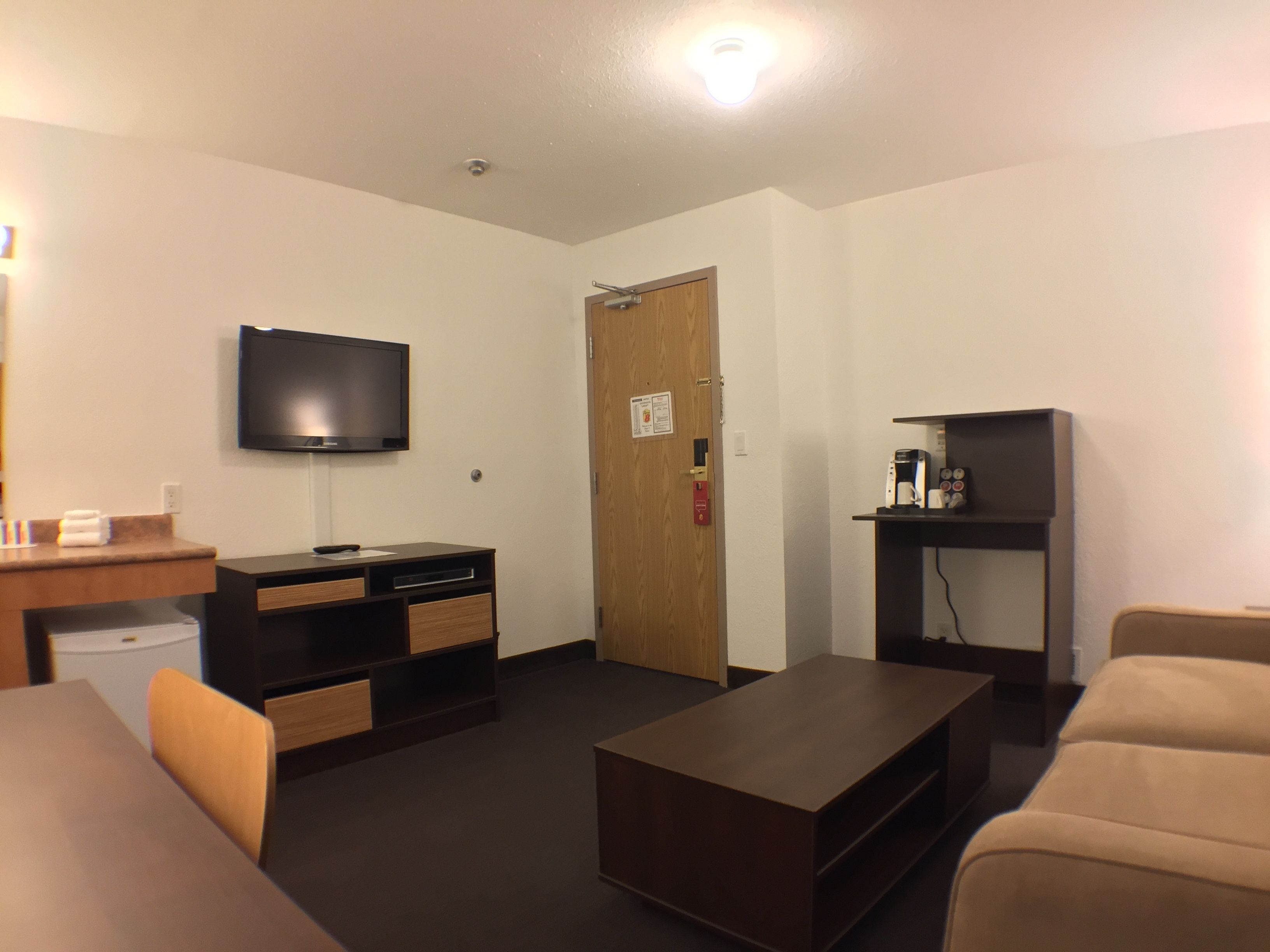 Una habitación de hotel compacta con una televisión de pantalla plana montada en la pared, una puerta de madera, una pequeña kitchenette con cafetera y un sofá.