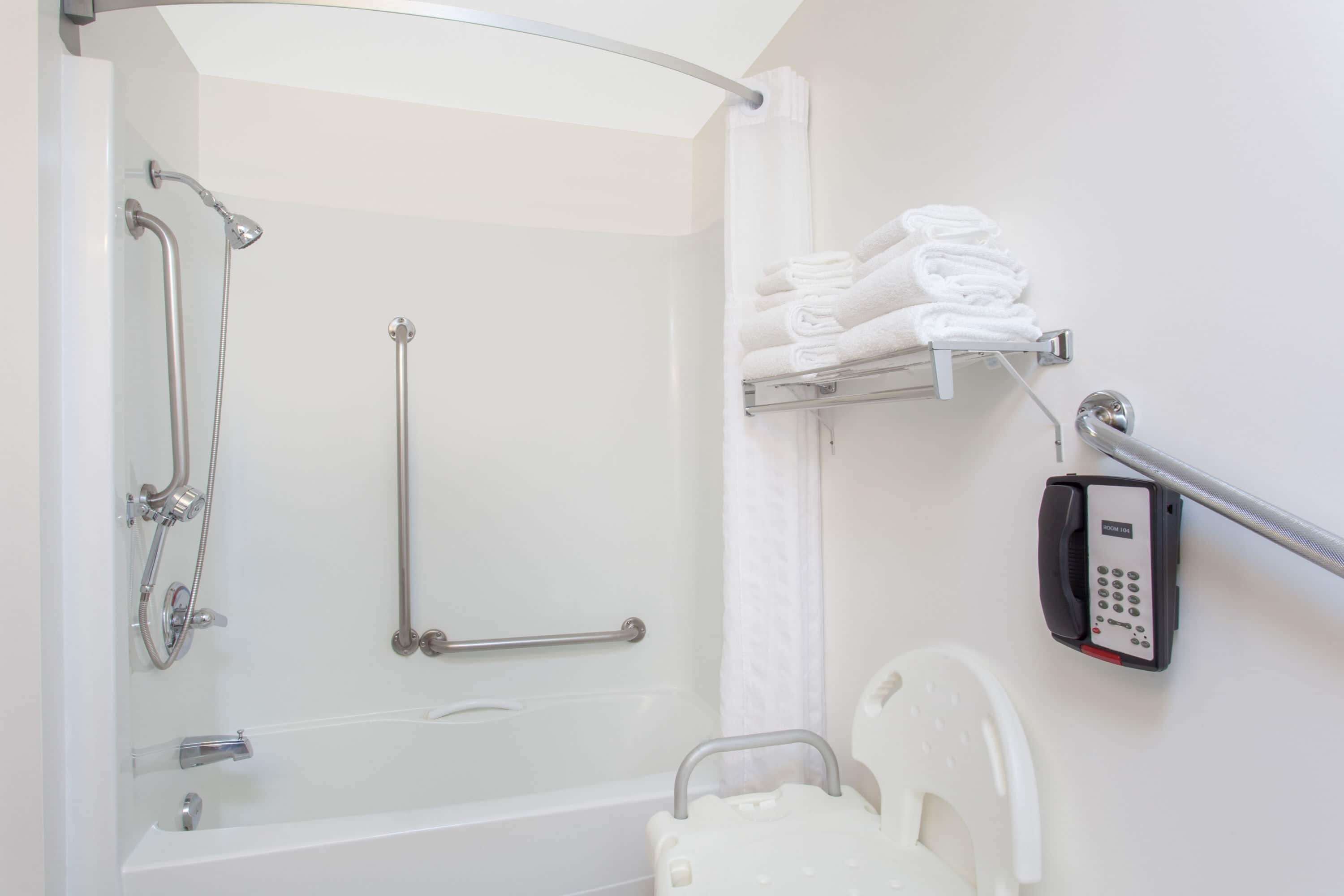 Ein Badezimmer mit Badewanne, einer Duschkabine mit Haltegriffen, einer Toilette mit Sicherheitsaufsatz und einem Handtuchhalter mit gefalteten Handtüchern.