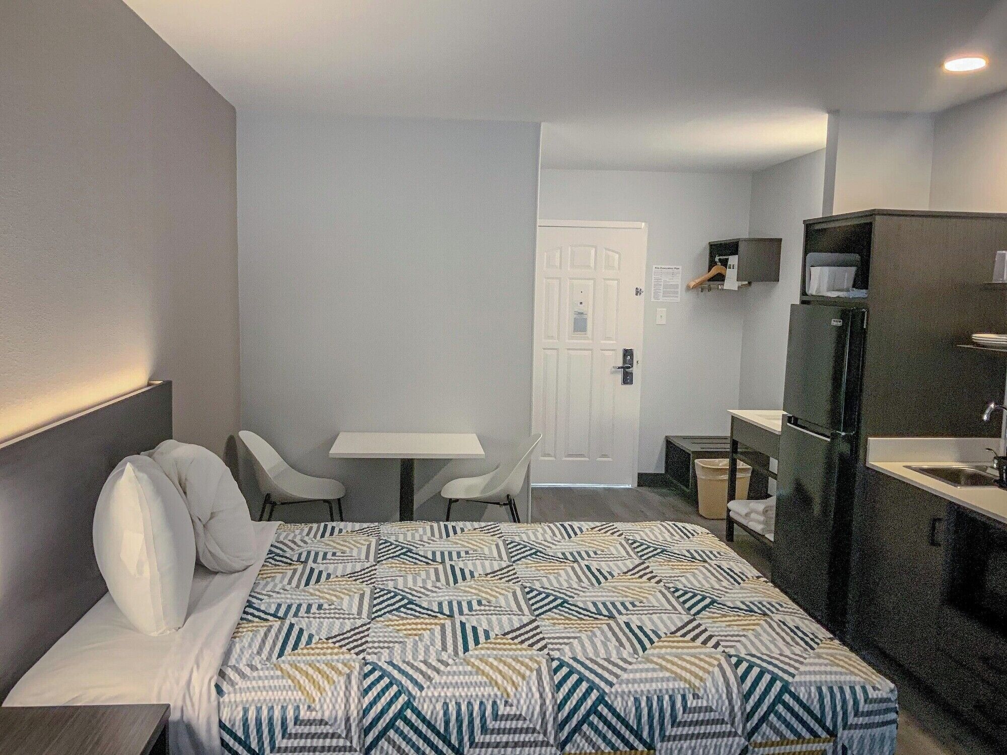 Una habitación de hotel con una cama, una mesita, dos sillas, una kitchenette y una puerta.