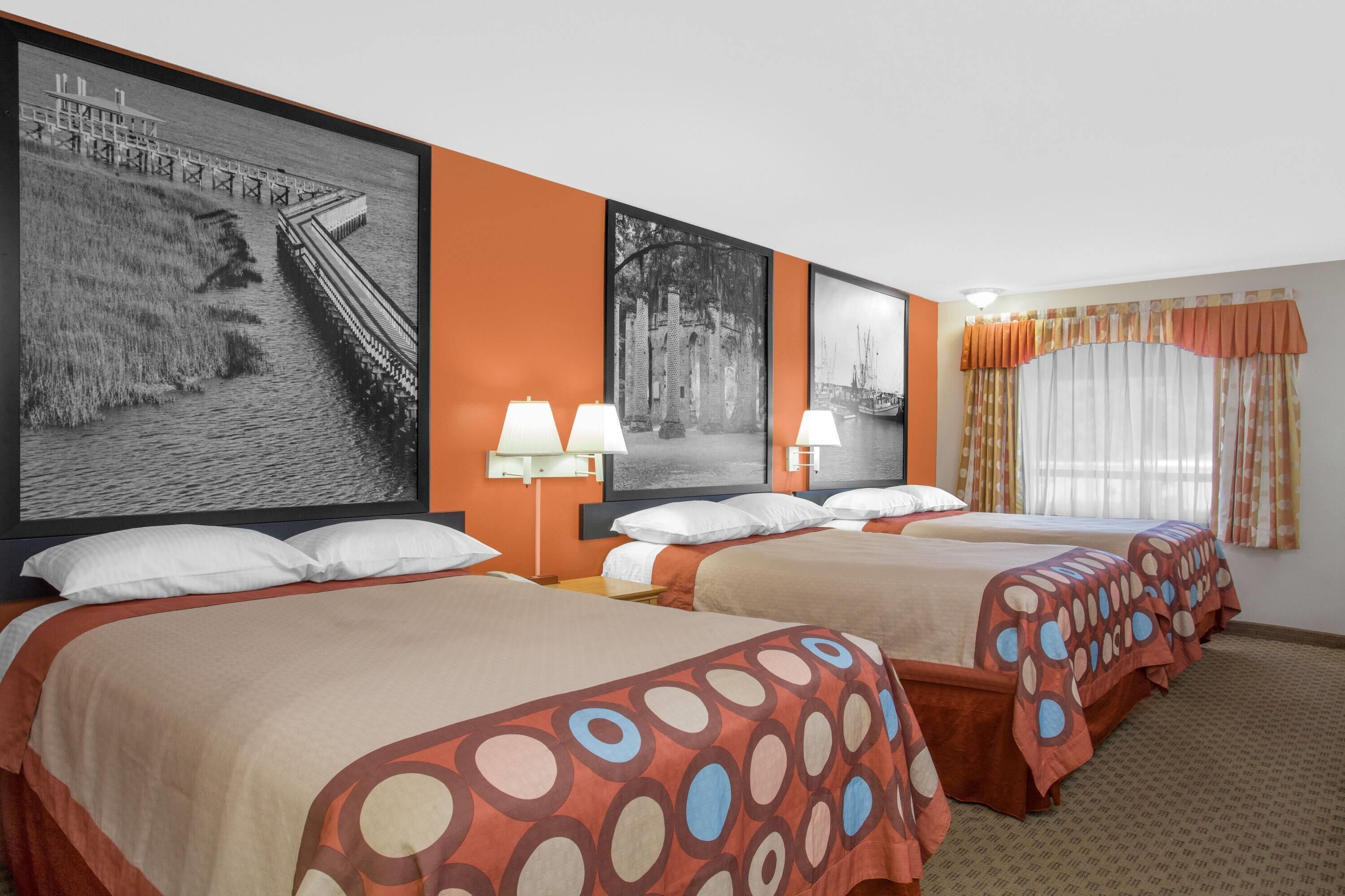 A hotel room with three beds, each with a patterned bedspread, a large framed photograph on the wall, and a window with curtains.
