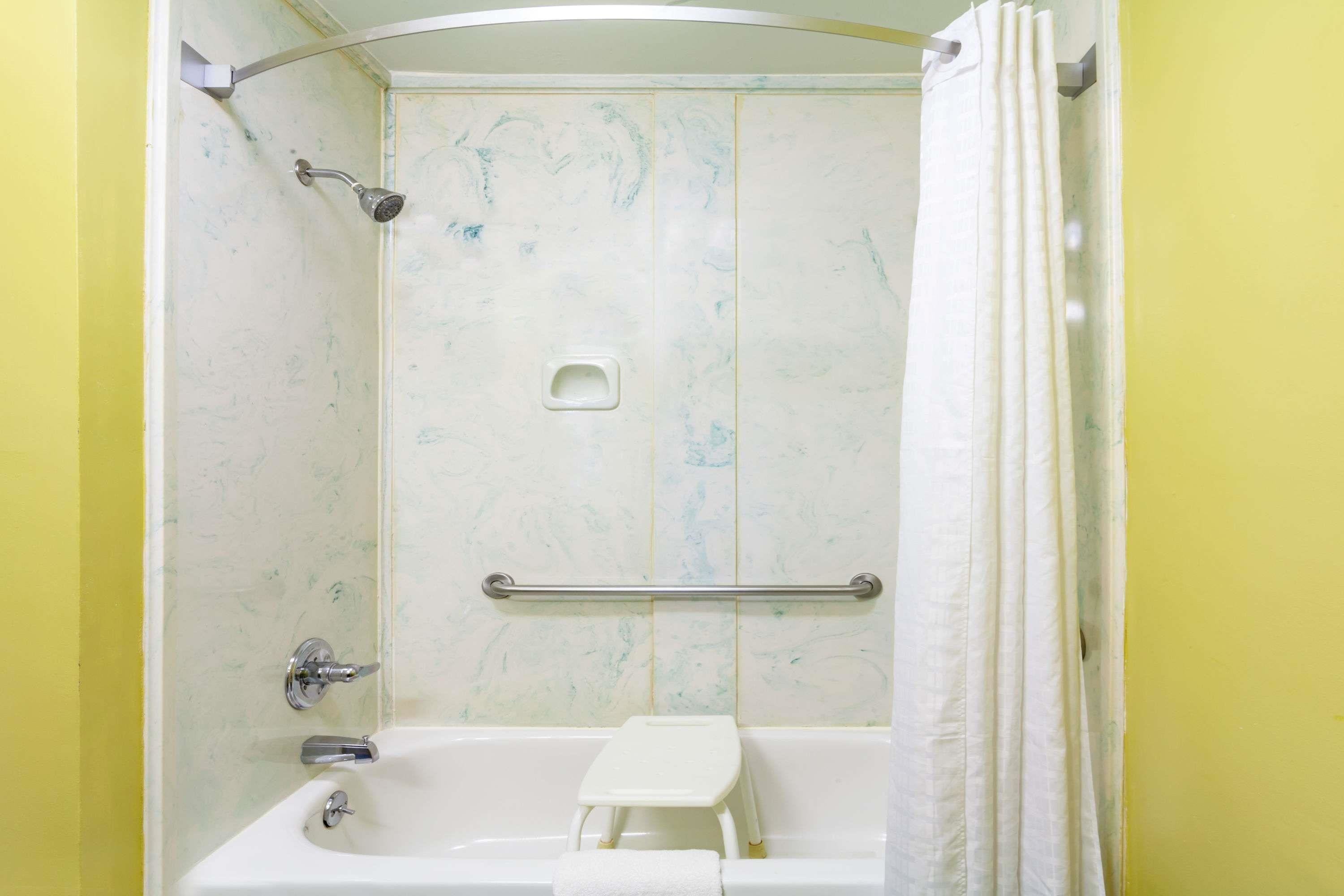 Ein Badezimmer mit einer weißen Badewanne, einer Dusche, einer Haltestange und einem kleinen weißen Hocker.
