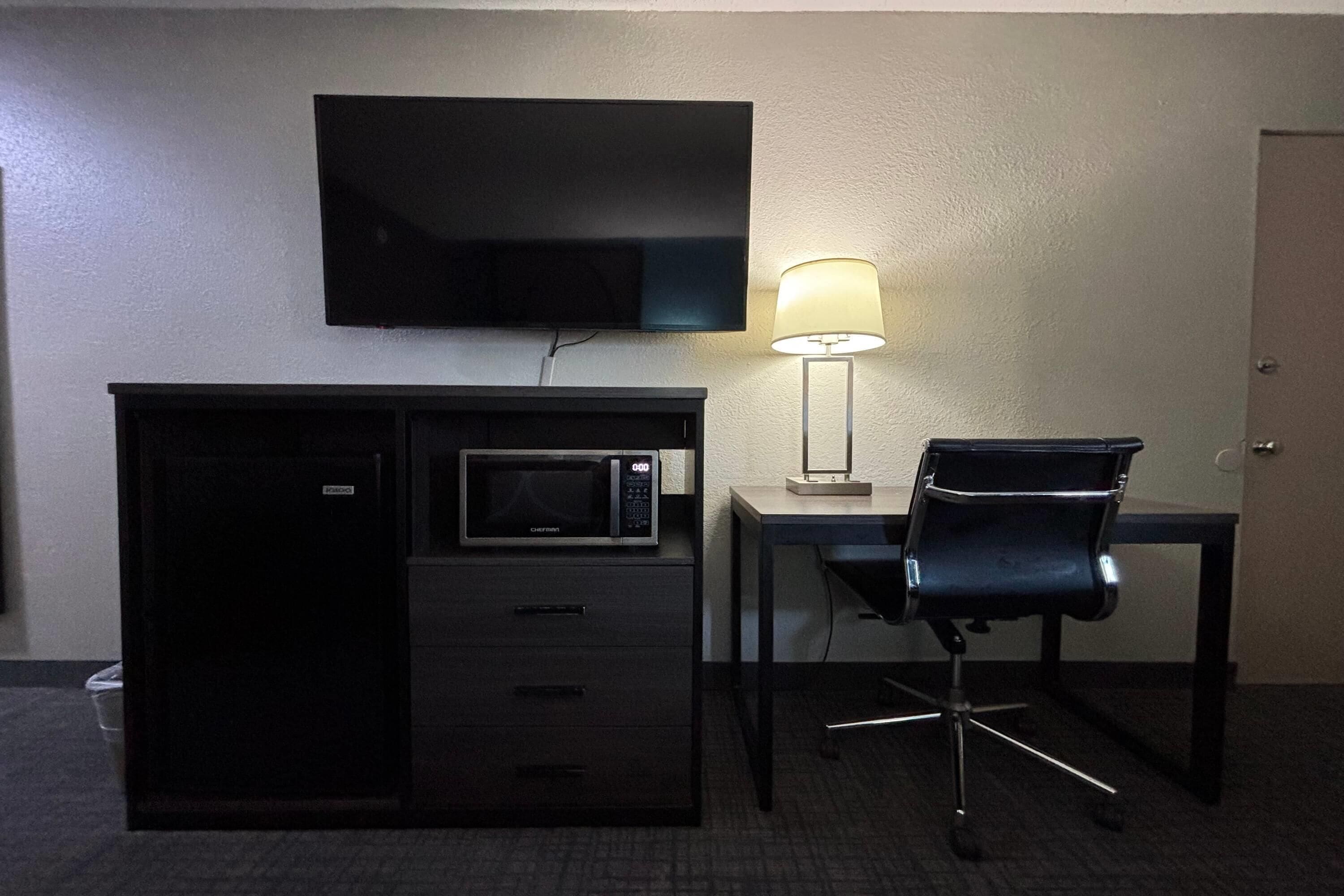 Ein kleines Hotelzimmer mit Schreibtisch, Mikrowelle, Fernseher, Stuhl und Lampe.
