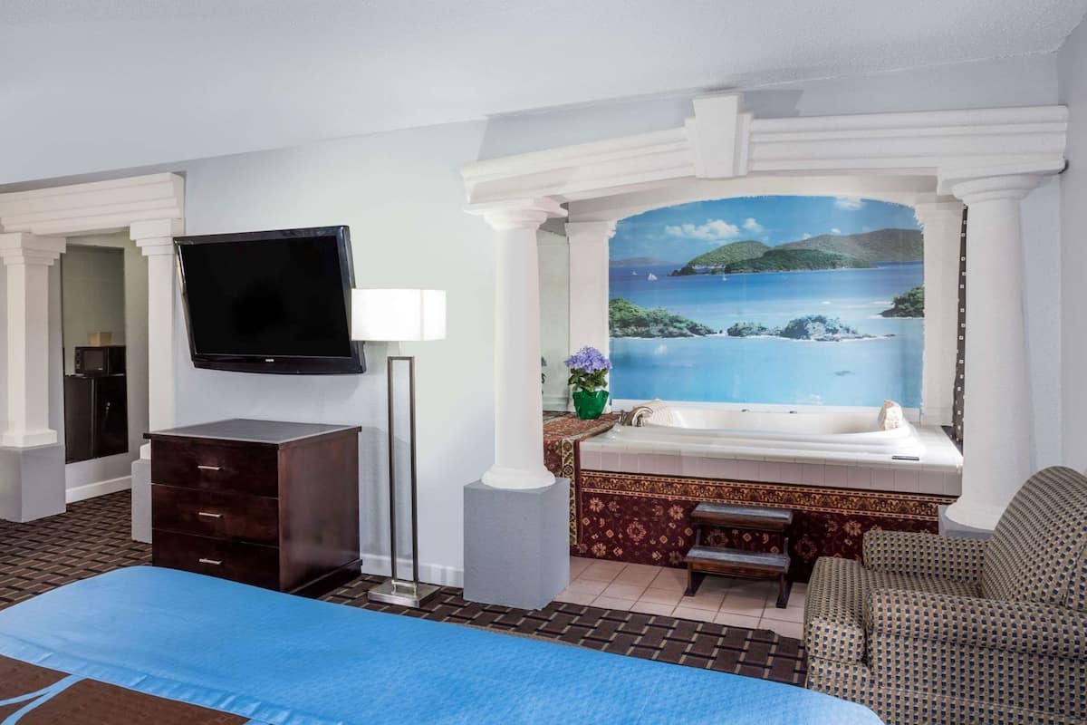 A hotel room with a bed, a TV, a desk, a chair, a window with a painting of a coastal scene, and a bathtub.