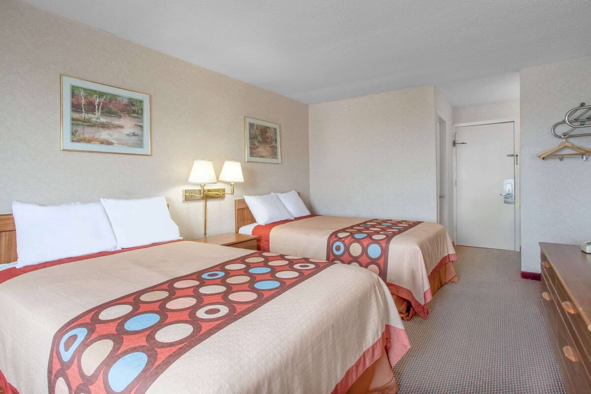 A hotel room with two beds, each with a patterned bedspread, a bedside table with a lamp, and a door with a coat rack.