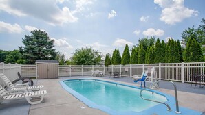 A rectangular swimming pool with a blue edge, surrounded by a white fence. There are lounge chairs and a table with chairs nearby.