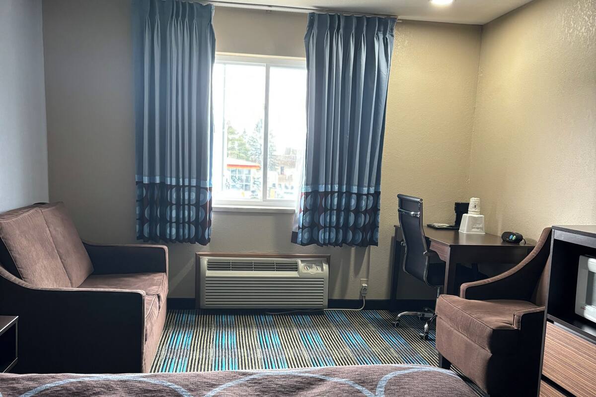 A hotel room with a bed, two chairs, a desk, an air conditioner, and a window with curtains.