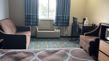 A hotel room with a bed, two chairs, a desk, an air conditioner, and a window with curtains.