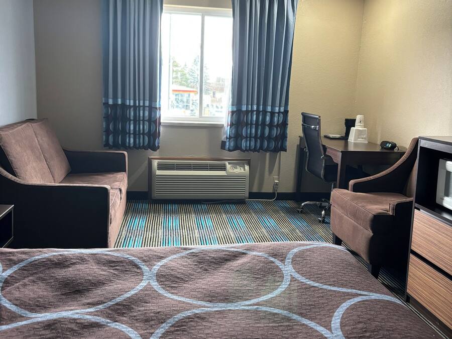 A hotel room with a bed, two chairs, a desk, an air conditioner, and a window with curtains.