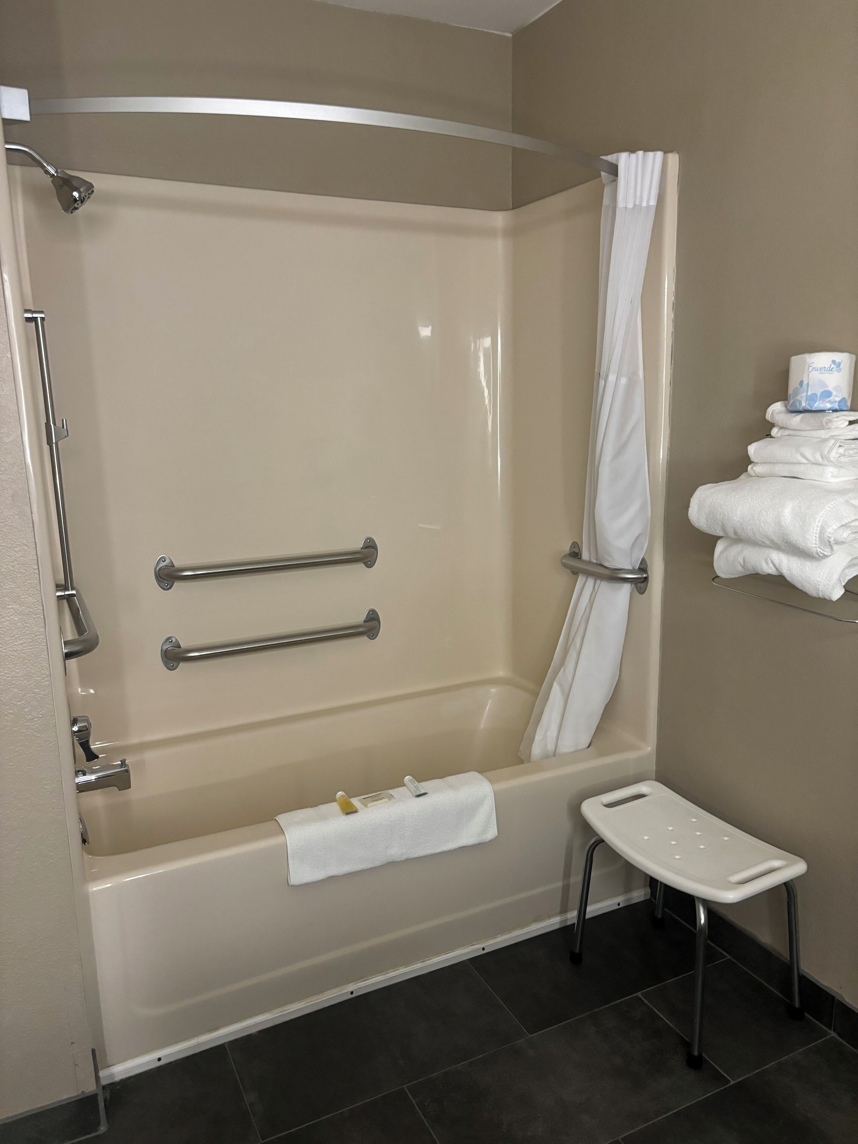 Un baño moderno con bañera, barras de apoyo, cortina de ducha, toallero y un pequeño taburete.