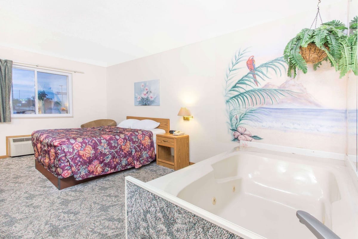 A hotel room with a bed, a nightstand, a lamp, a painting, a window, and a bathtub.