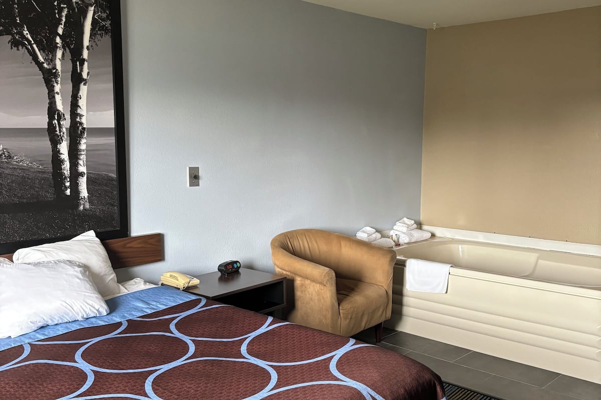 A hotel room with a bed, a chair, a large painting, and a bathtub.