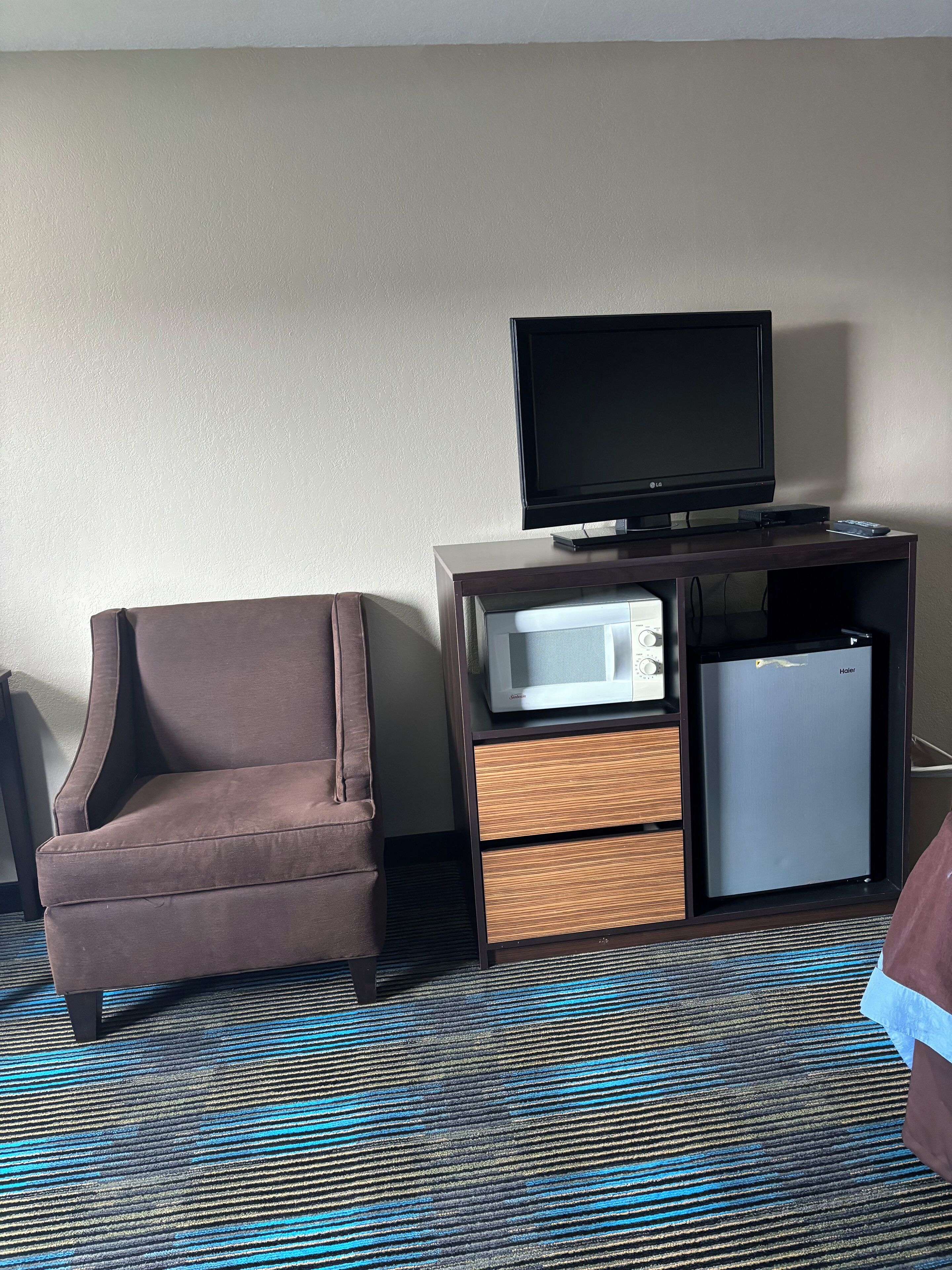 A hotel room with a brown armchair, a flat-screen TV, a microwave, and a mini-fridge.