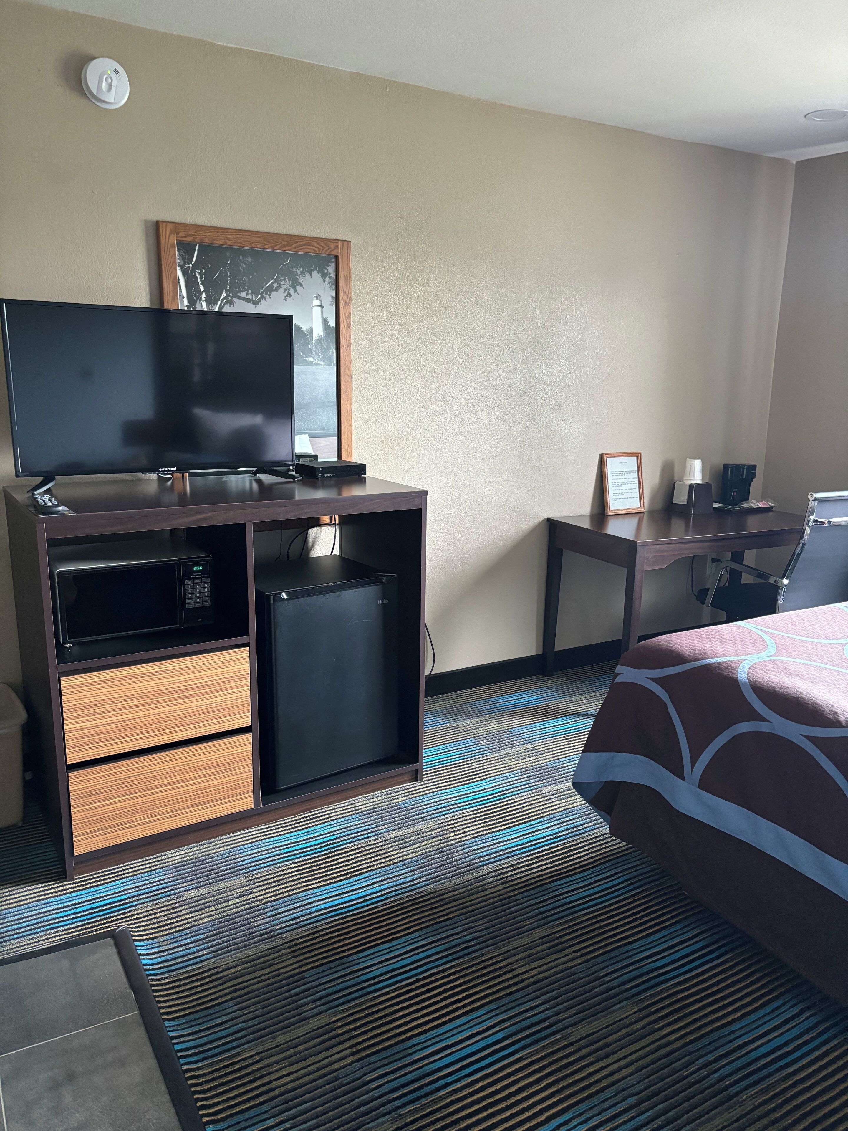 A hotel room with a flat-screen TV, a small refrigerator, a desk with a computer, a bed with a patterned bedspread, and a window with a view of trees and a lighthouse.