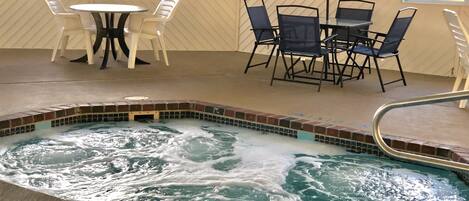 A hot tub with a tiled edge, surrounded by a pool. There are chairs and a table with an umbrella.