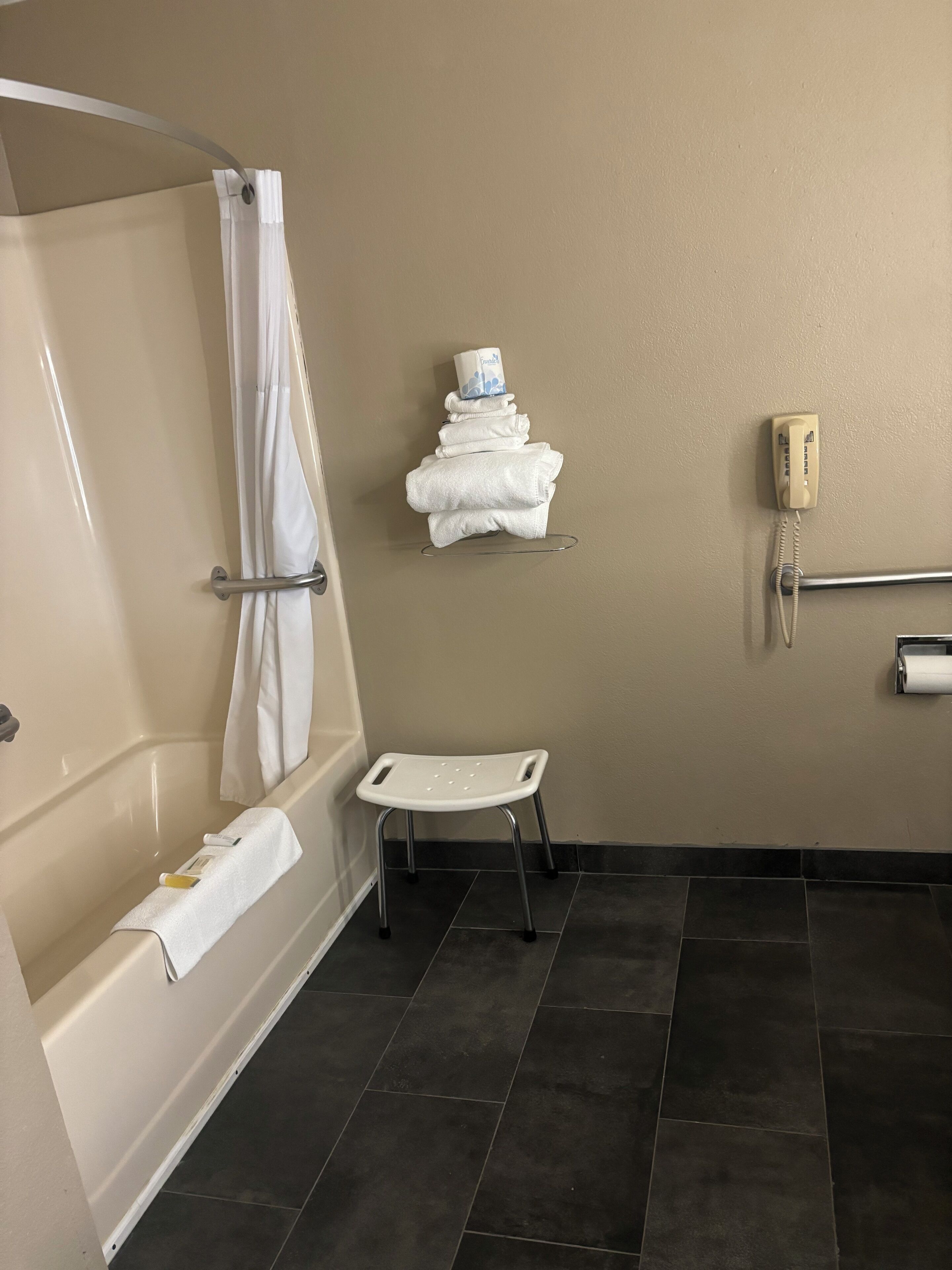 A bathroom with a bathtub, a shower curtain, a towel rack with folded towels, a telephone, and a grab bar.