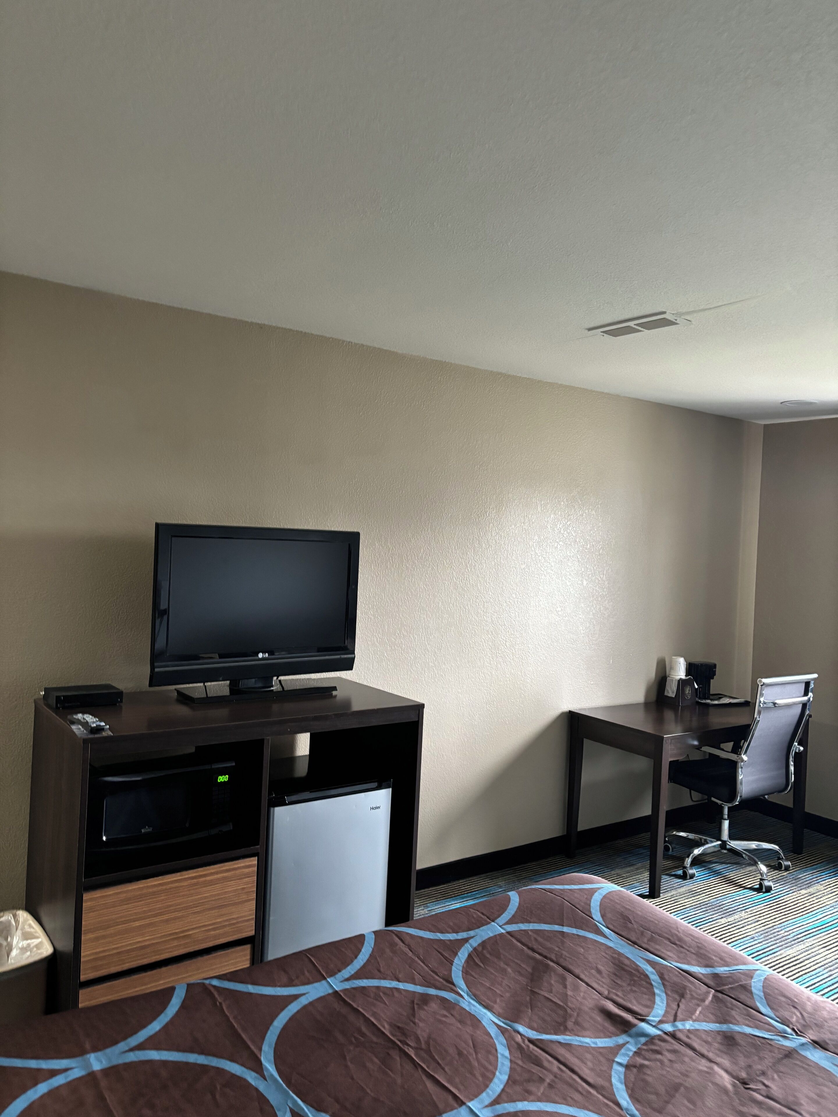 A hotel room with a bed, a desk with a television, a small refrigerator, and a chair.