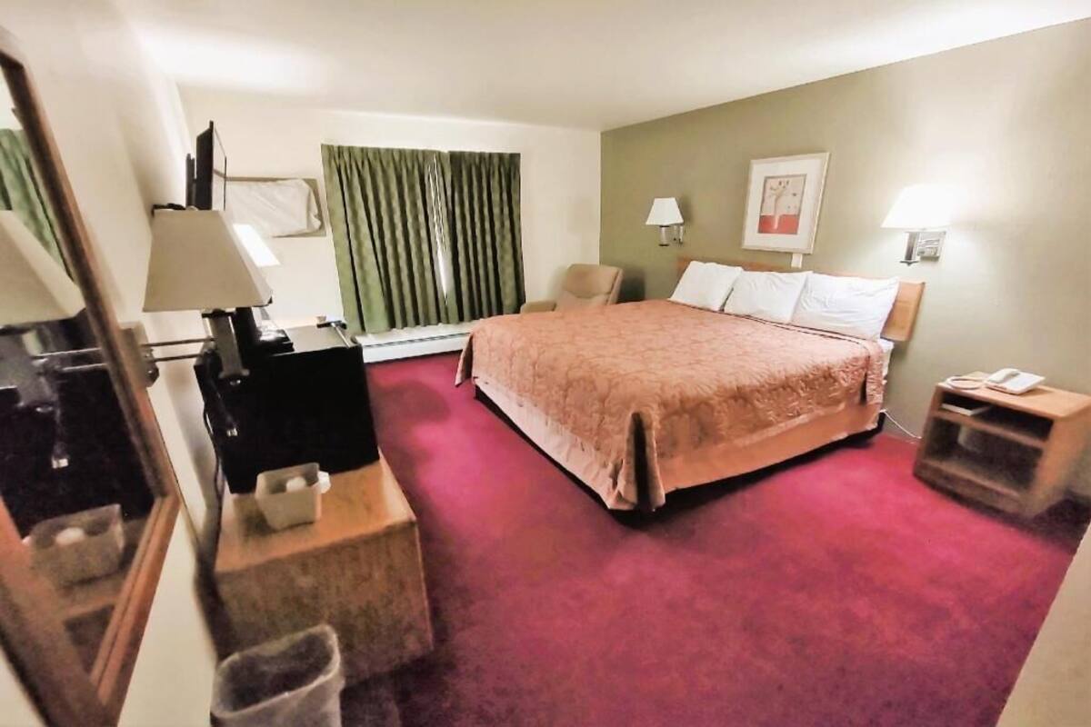 A hotel room with a bed, a desk, a chair, a TV, a lamp, a trash bin, and a window with curtains.