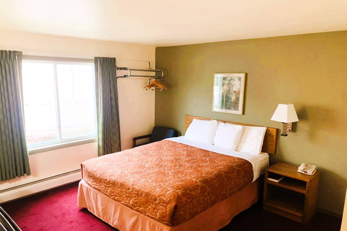 A hotel room with a large bed, a bedside table with a lamp, a small table with a phone, and a window with curtains.