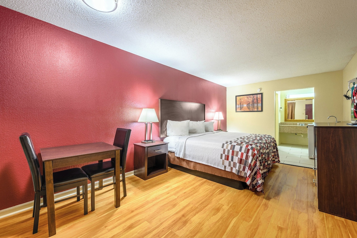 A hotel room with a bed, a desk with a chair, a bedside table with lamps, and a mirror.