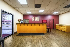 A reception area with a wooden reception desk, a red door, and a window overlooking a parking lot. - Red Roof Inn Sylacauga (Sylacauga)
