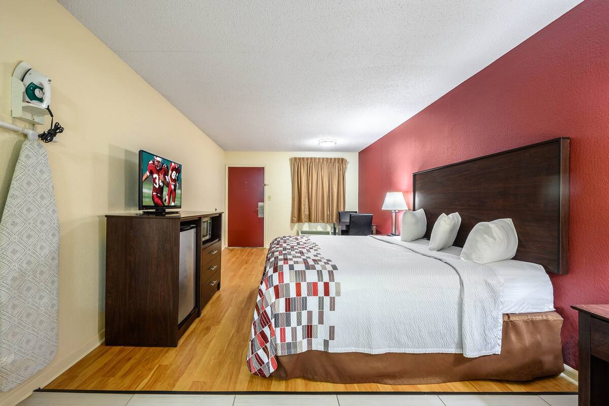 A hotel room with a large bed, a desk with a television, a lamp, and a red accent wall.