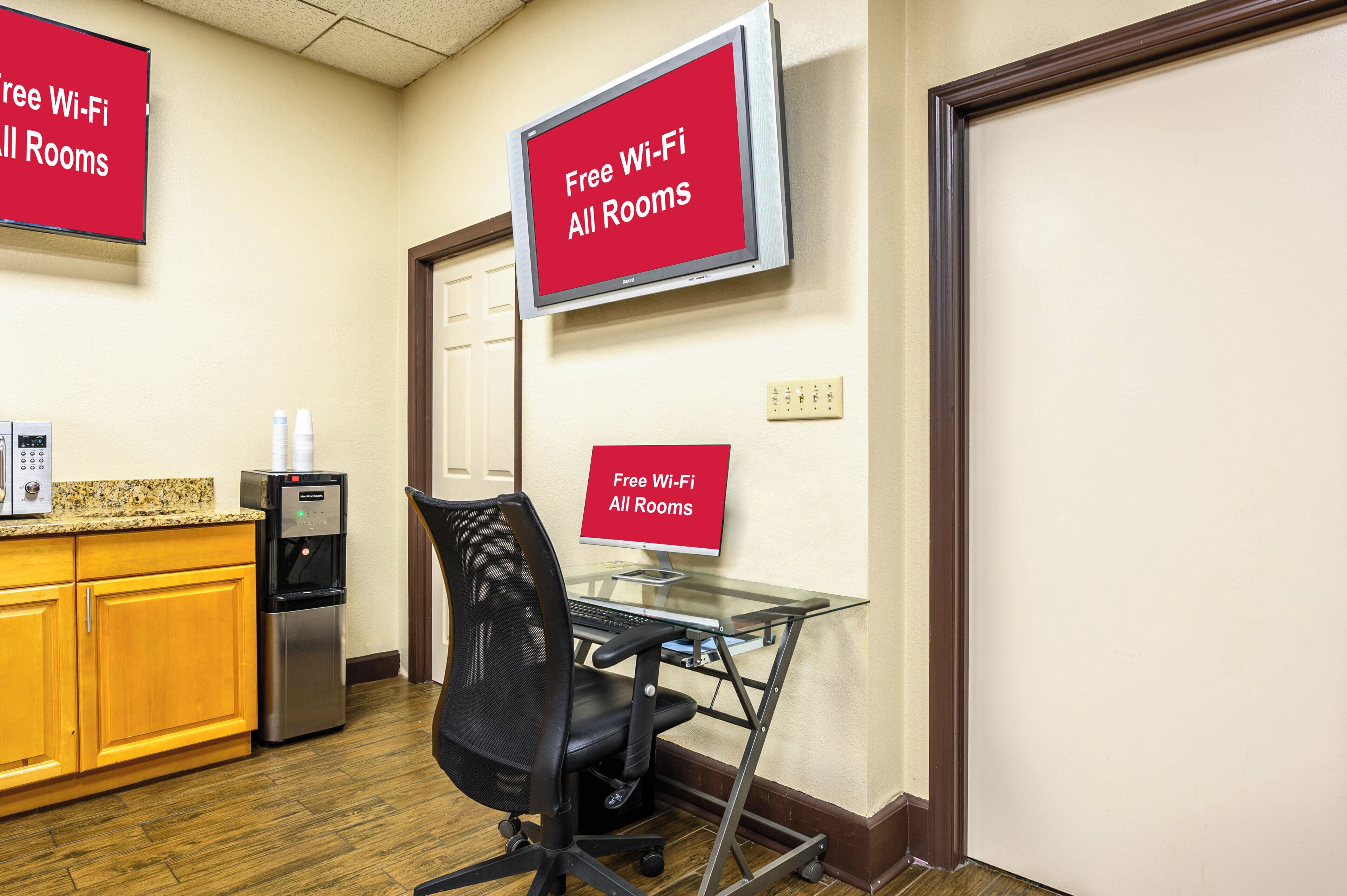 A hotel room with a kitchenette, a water dispenser, a glass desk with a chair, and signs indicating free Wi-Fi in all rooms.