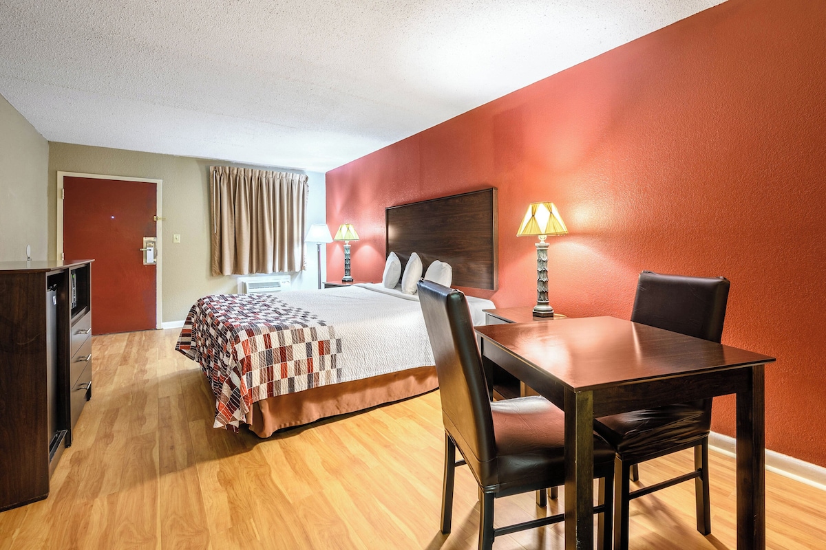 A hotel room with a bed, a desk with a chair, a lamp, and a door.