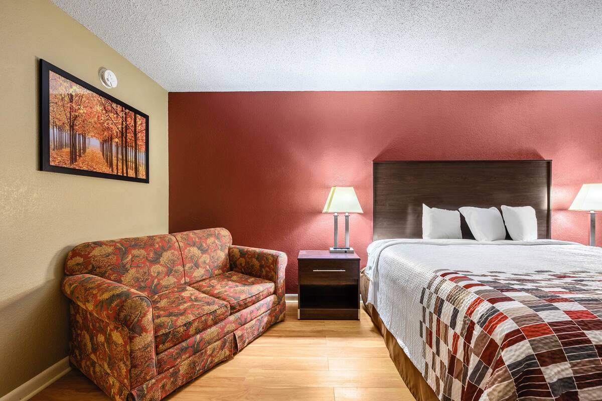 A hotel room with a bed, a nightstand with a lamp, a sofa, and a painting on the wall.