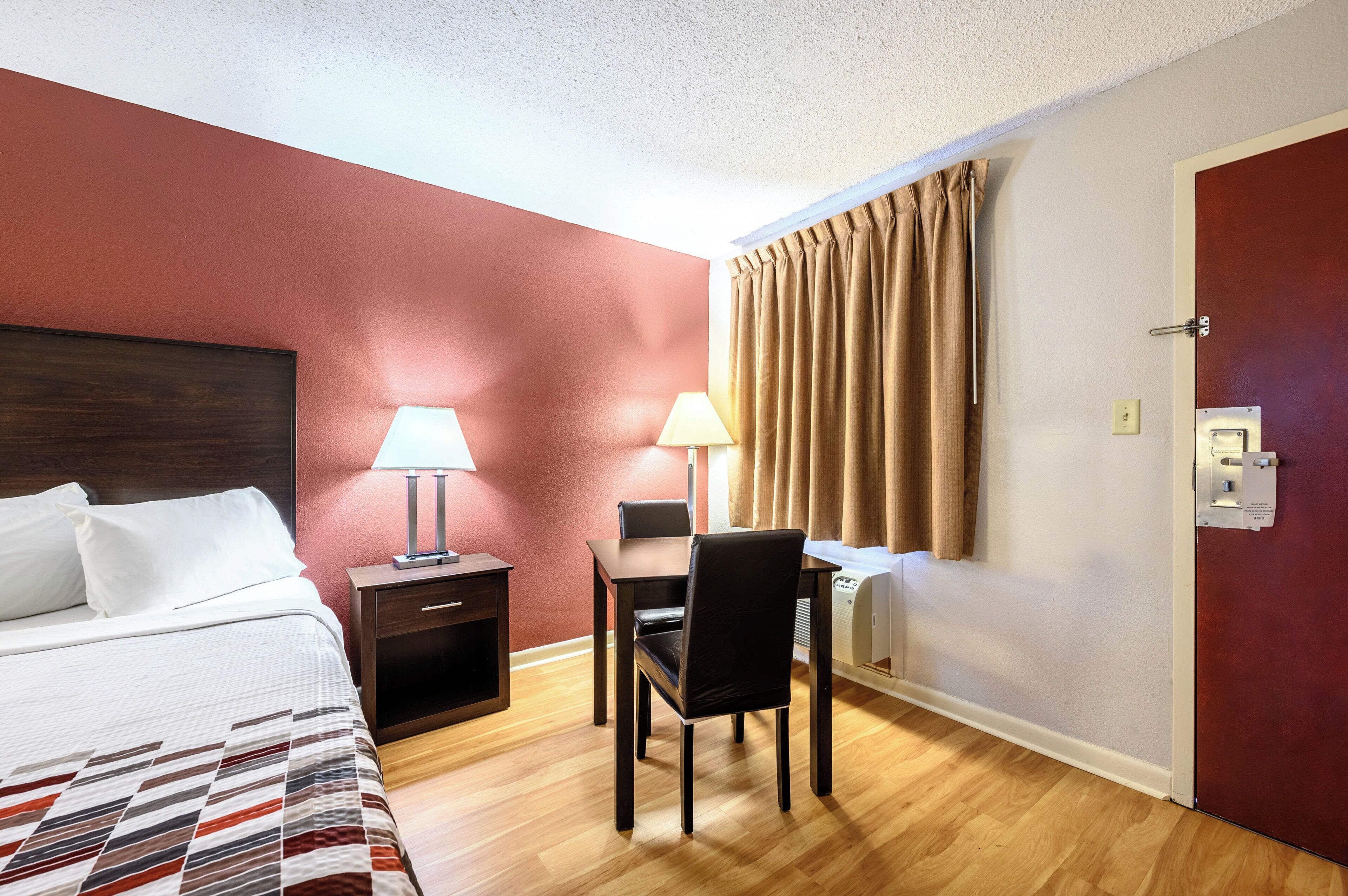 A hotel room with a bed, a desk, a chair, a lamp, and a door.