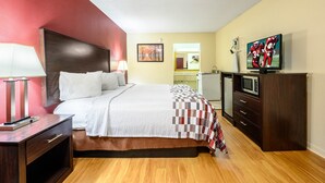 A hotel room with a bed, bedside table, lamp, TV, and a dresser. - Red Roof Inn Sylacauga (Sylacauga)