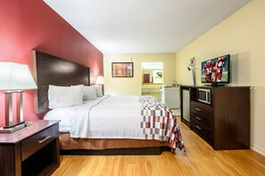 A hotel room with a bed, bedside table, lamp, TV, and a dresser. - Red Roof Inn Sylacauga (Sylacauga)