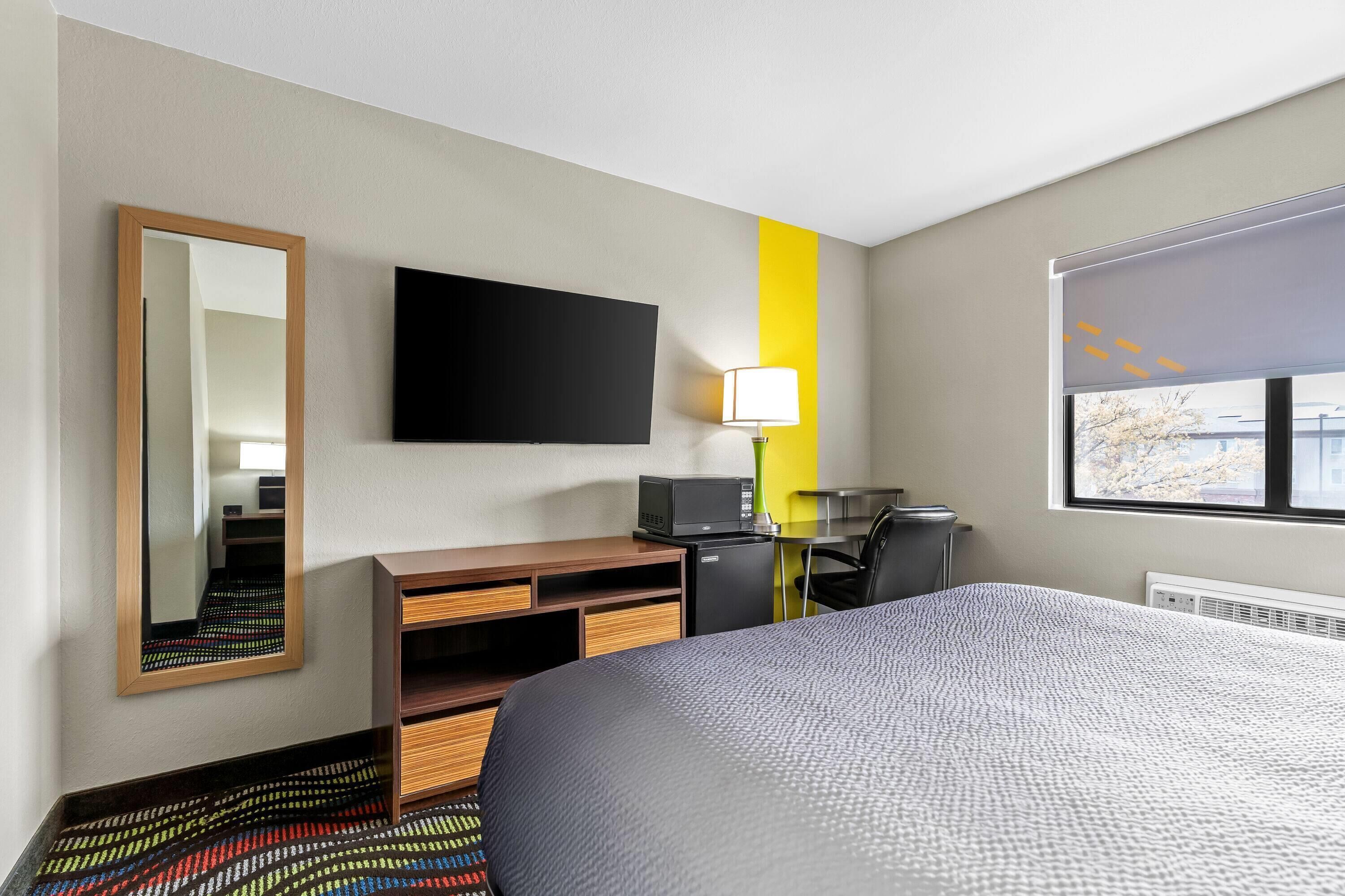 Habitación de hotel con cama, escritorio con computadora, espejo, lámpara y ventana.