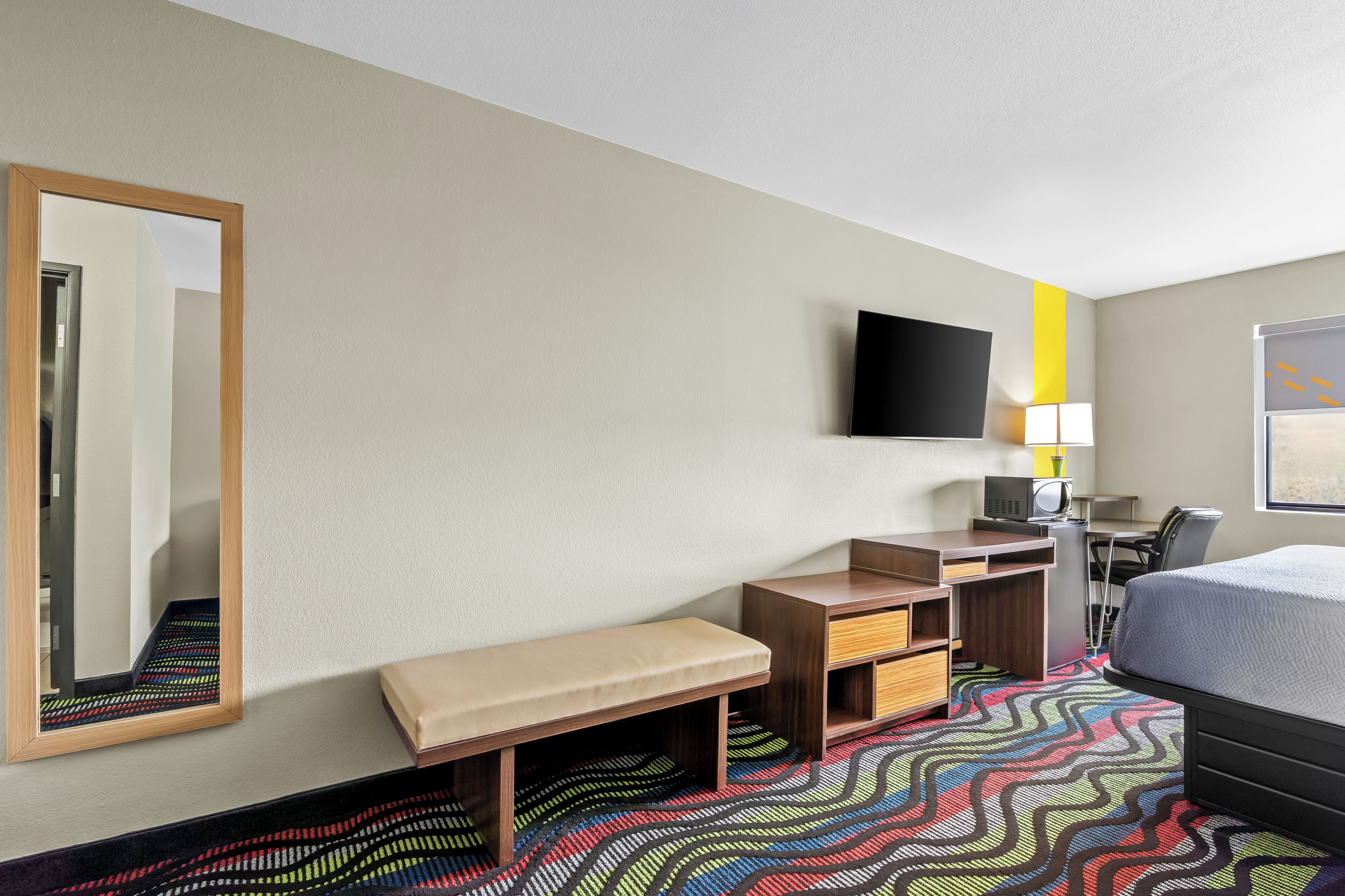 Habitación de hotel con cama, escritorio con silla, banco, televisor y espejo.