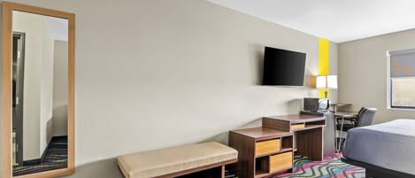 Habitación de hotel con cama, escritorio con silla, banco, televisor y espejo.