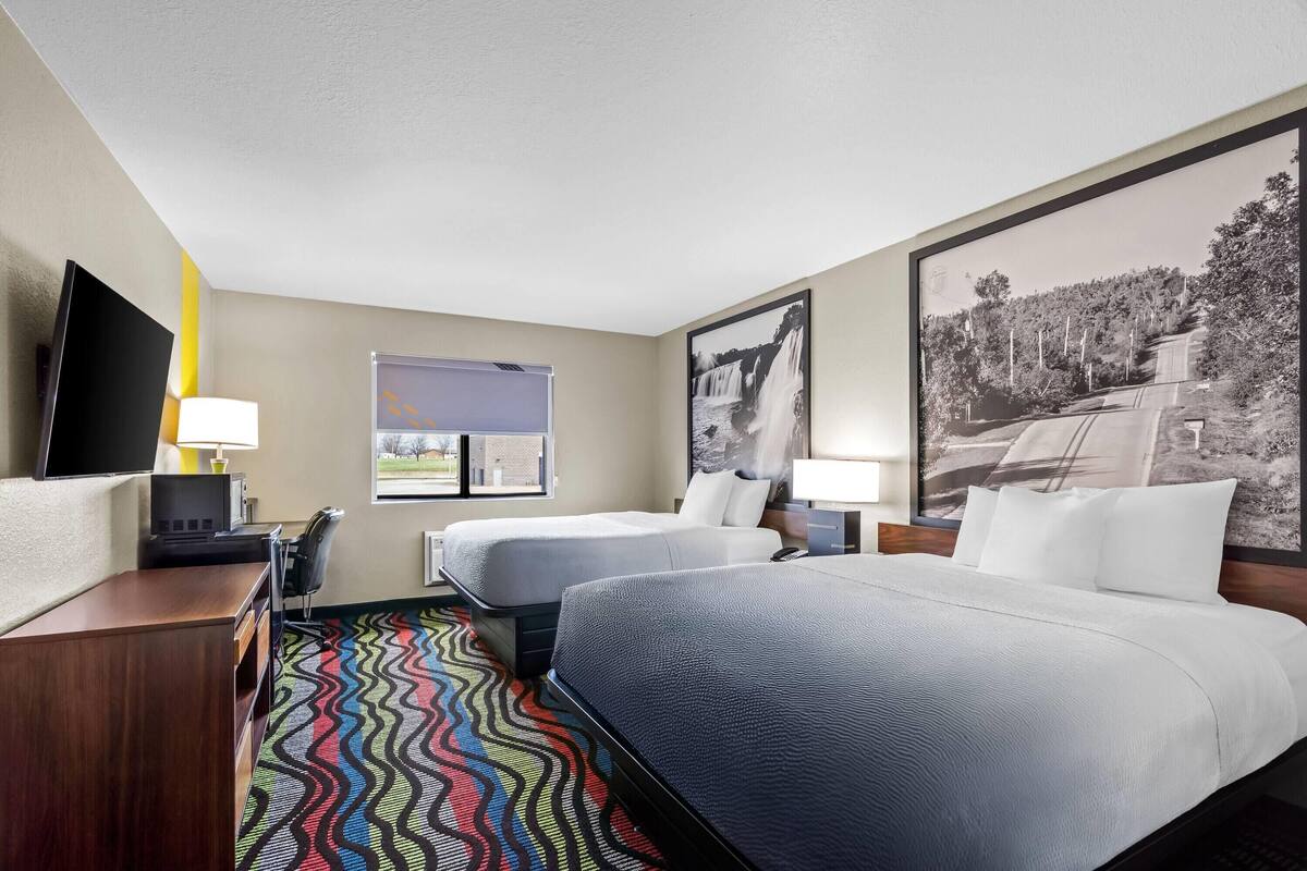 Une chambre d’hôtel avec deux lits, une télévision, un bureau, une lampe, une fenêtre et une moquette colorée.