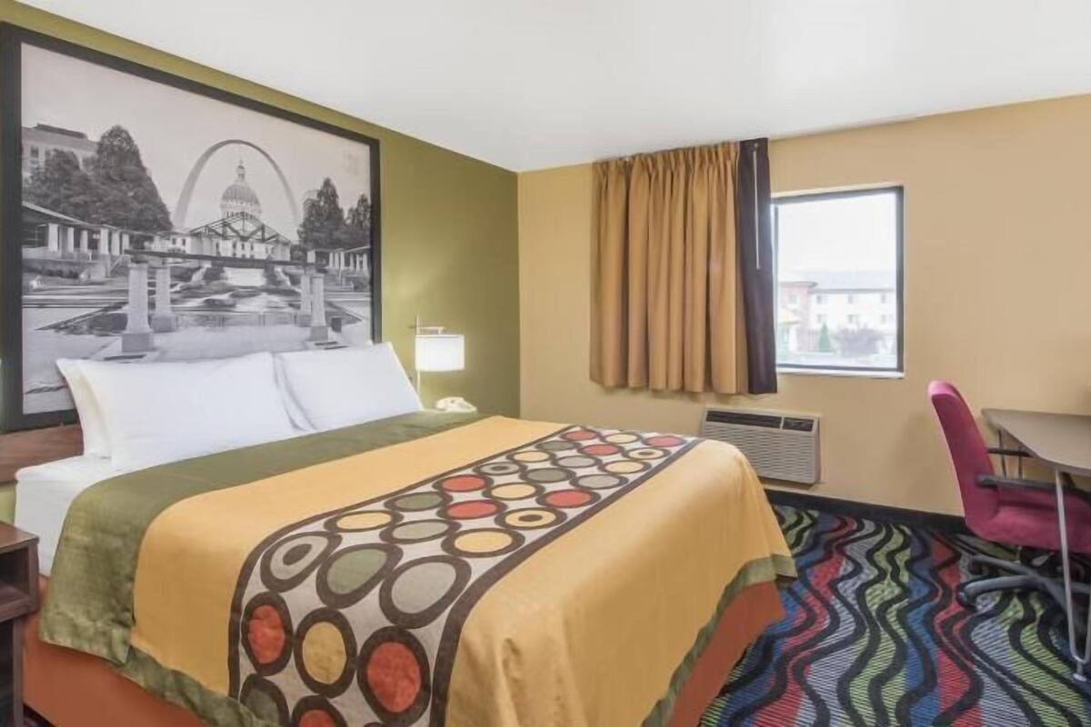 Une chambre d’hôtel comprenant un lit, un bureau, une chaise, une fenêtre avec des rideaux, une lampe fixée au mur et un grand tableau encadré.
