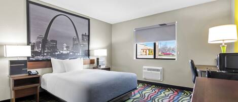 A hotel room with a bed, nightstands, a desk, a television, a colorful rug, a cityscape poster, and a window with a view of a building.