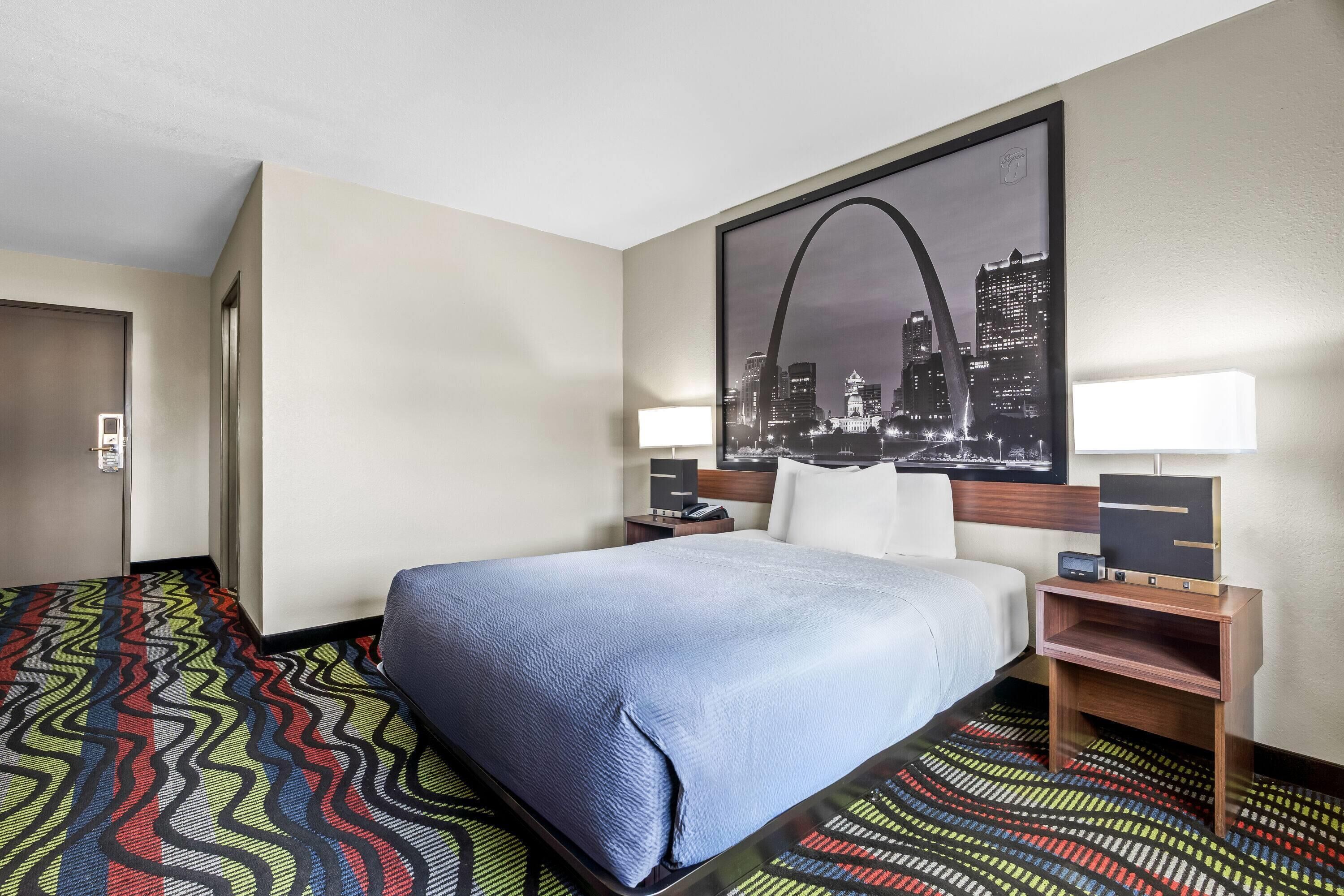 A hotel room with a bed, a nightstand, a lamp, a desk, a chair, a patterned carpet, and a cityscape painting on the wall.