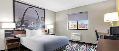 Quarto de hotel com cama, duas mesas de cabeceira, escrivaninha, cadeira, tapete colorido, pôster com vista urbana, janela e abajur.