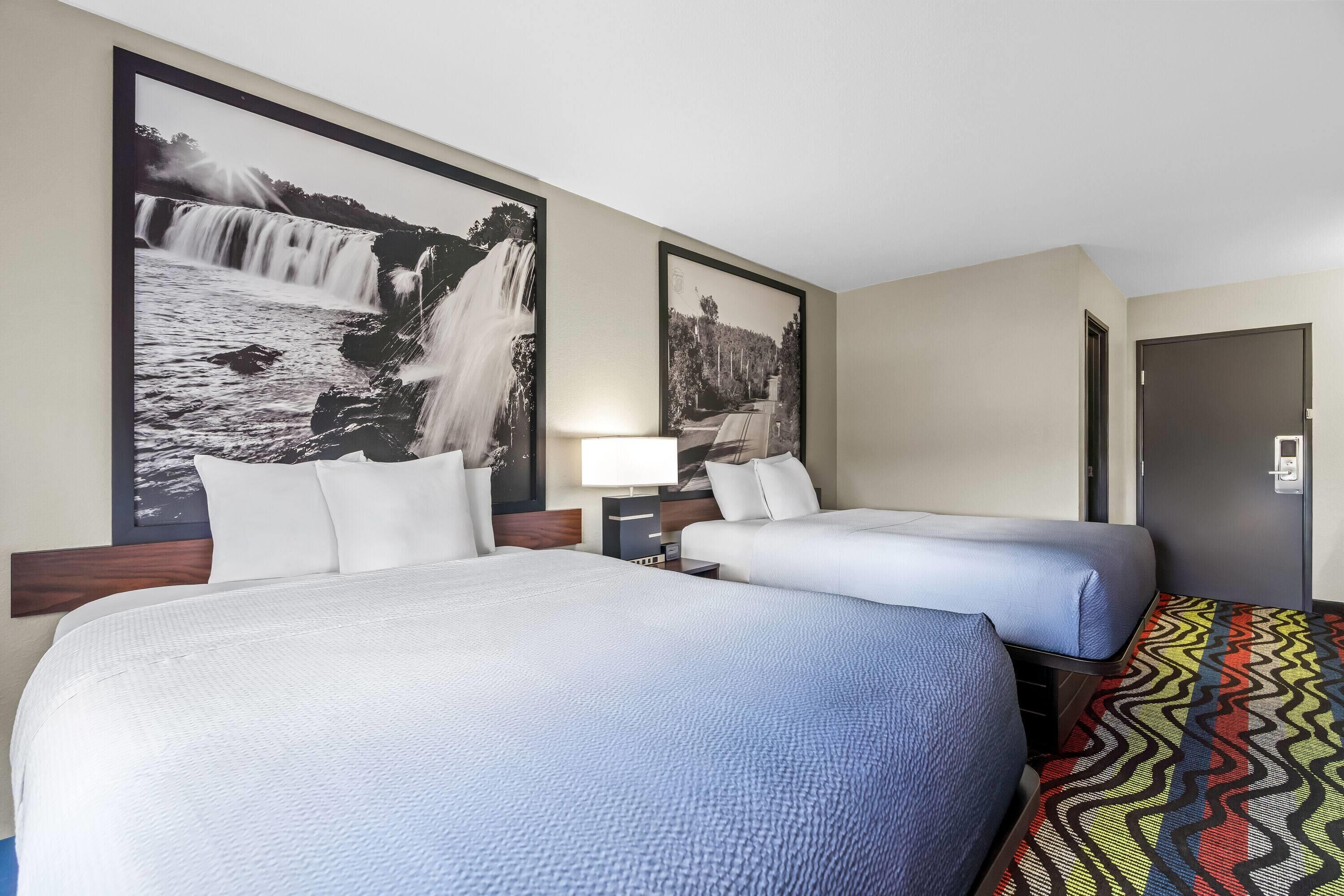 A hotel room with two beds, a large wall-mounted picture of a waterfall, a bedside table with a lamp, and a door.