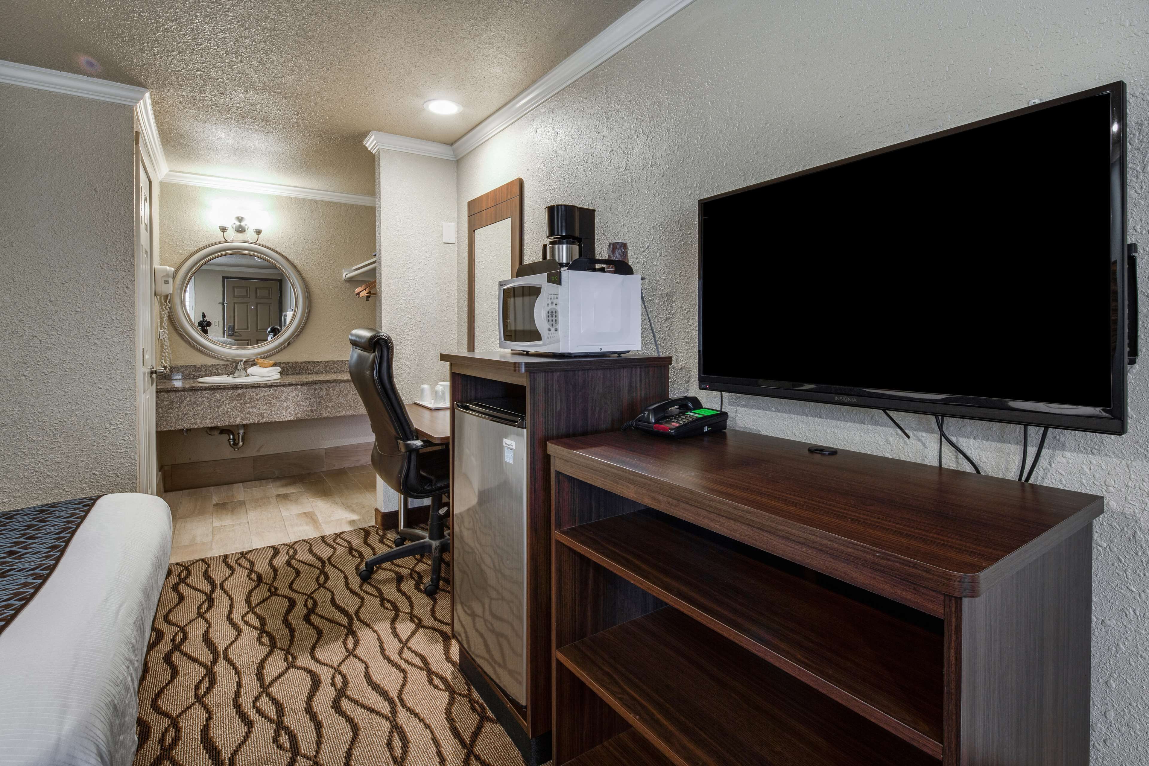 Habitación de hotel con cama, escritorio con microondas, cafetera, televisión y baño con lavabo y espejo.