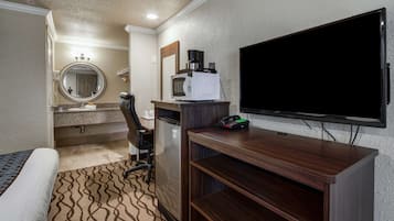Habitación de hotel con cama, escritorio con microondas, cafetera, televisión y baño con lavabo y espejo.
