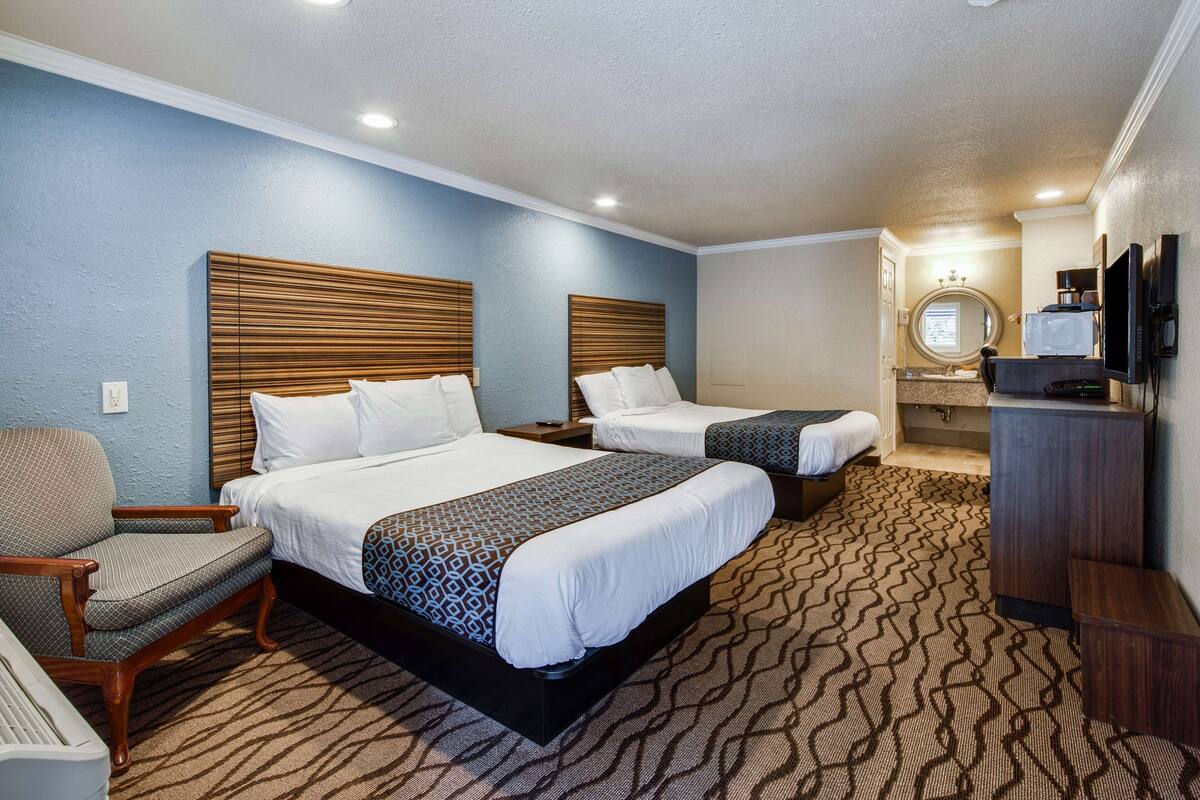 A hotel room with two beds, a chair, a desk, a television, and a bathroom with a mirror and sink.