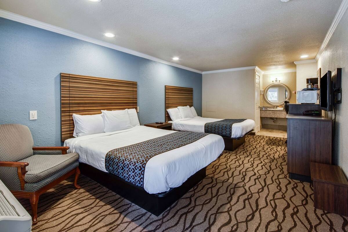 A hotel room with two beds, a chair, a desk, a television, and a bathroom with a mirror.