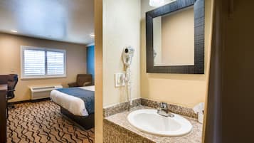 A hotel room with a bed, a desk, a chair, a window with blinds, a hairdryer, a sink, and a mirror.